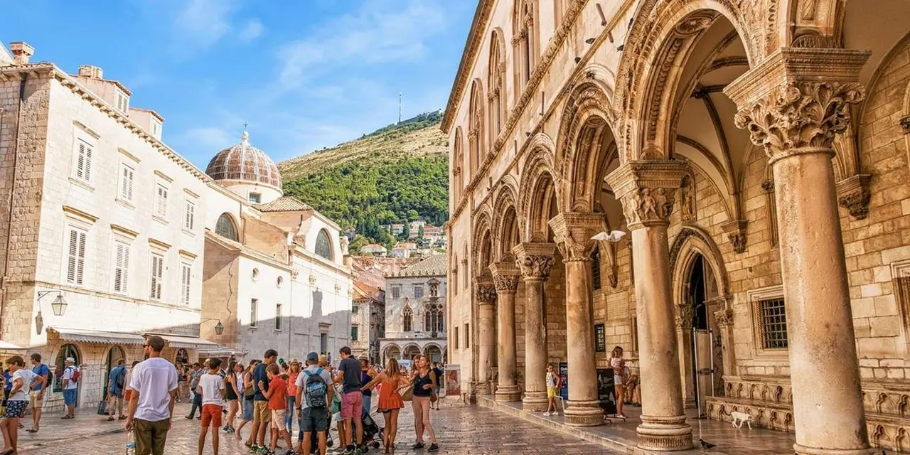 The arcaded stone facade of the Rector's Palace with visitors in the street and green hills behind