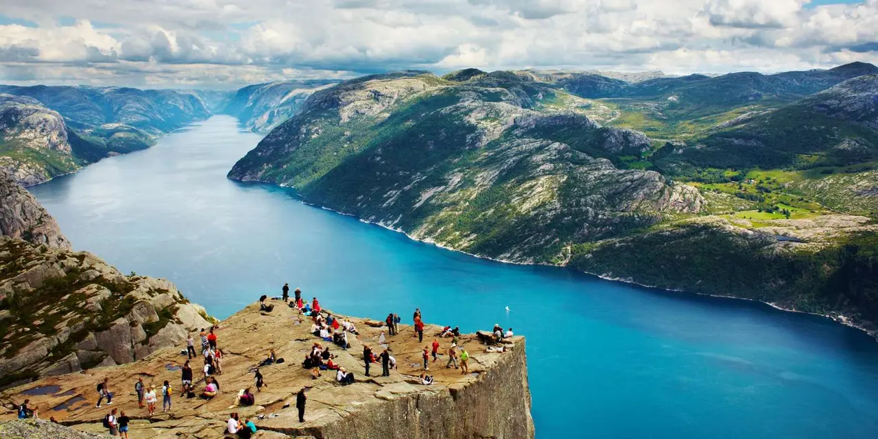 Hikers on the flat Pulpit Rock plateau high above a bright blue fjord, with green mountains beyond