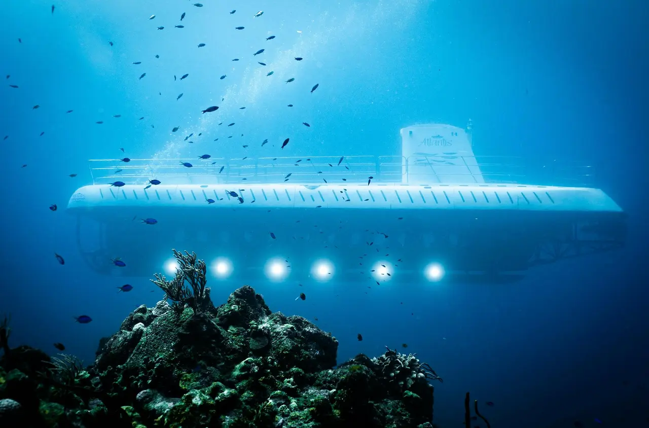 The white Atlantis submarine lit from below above a coral outcrop, surrounded by schools of small fish in deep blue water