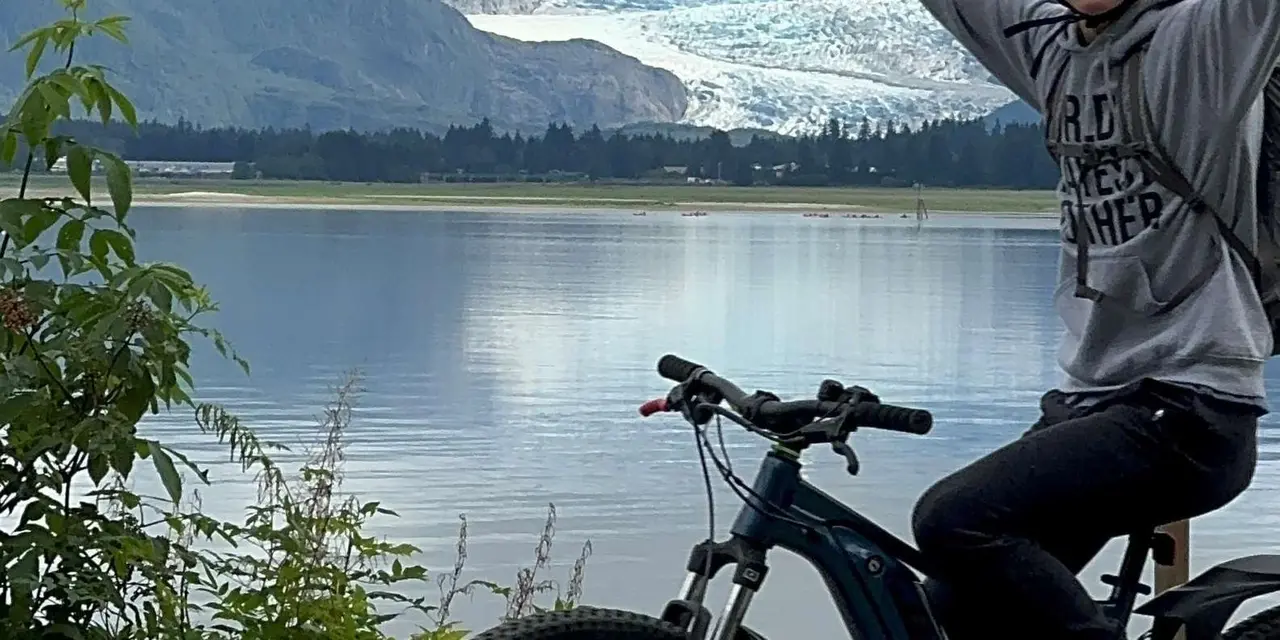 A mountain bike and a rider in a grey hoodie with arms raised beside a still lake, with the glacier across the water and green forest on the left