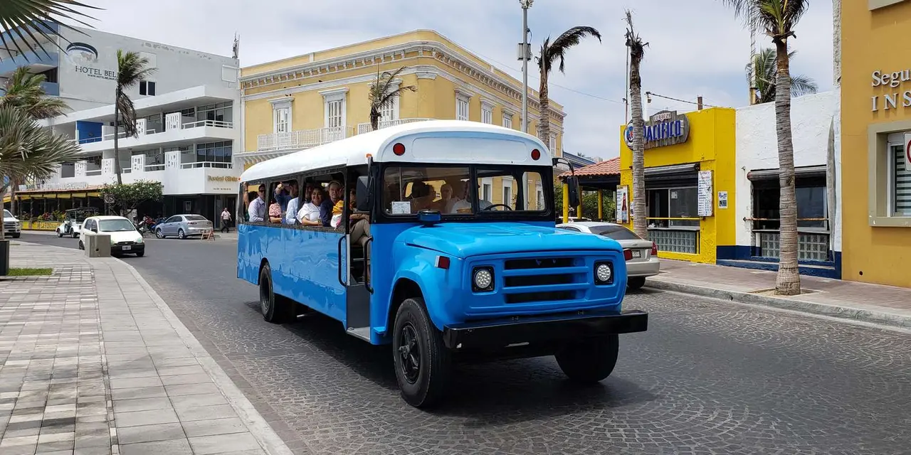 A blue city bus with passengers aboard driving past yellow and white buildings on a palm-lined street