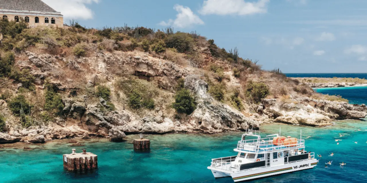 A white two-deck catamaran moored in a turquoise cove below a rocky, scrub-covered headland