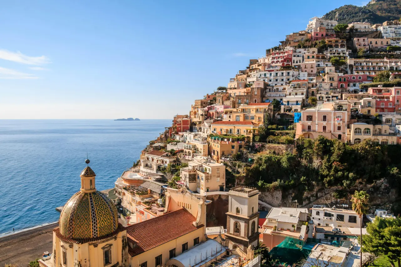 Positano's pastel houses stacked down a cliff above the sea, with a tiled church dome in the foreground