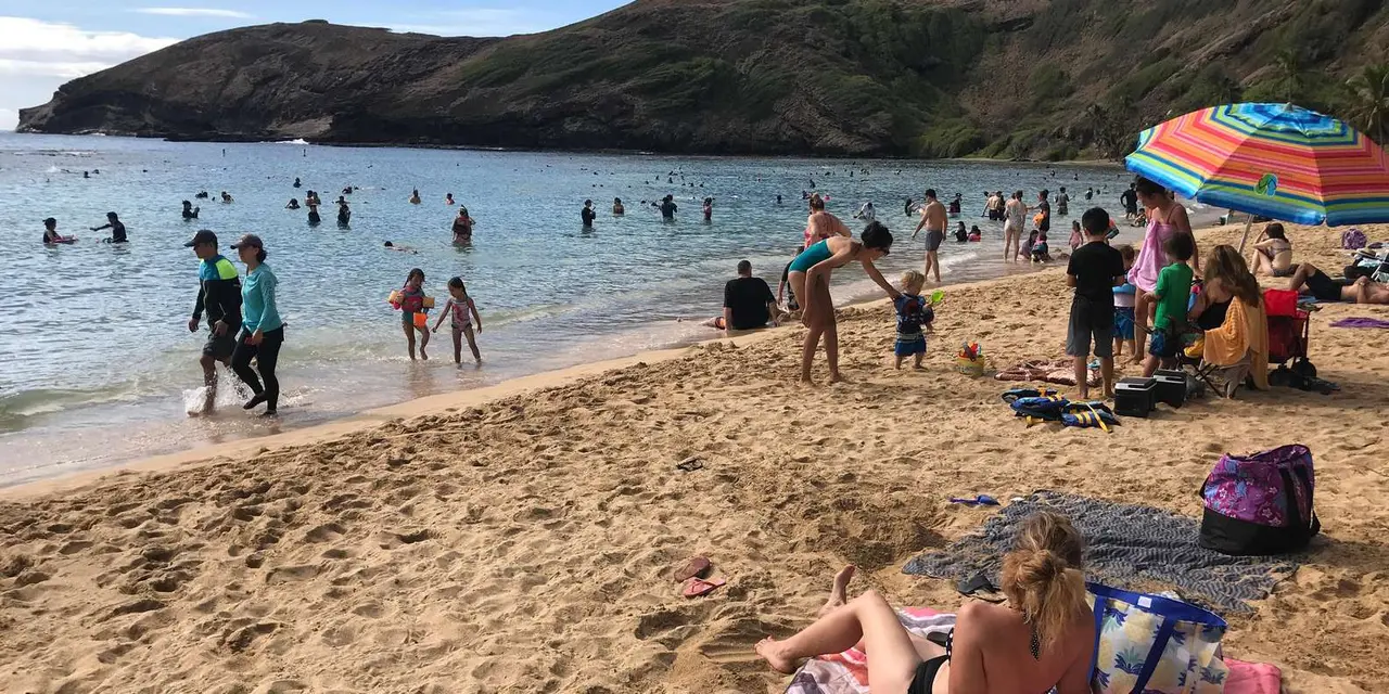Swimmers in shallow turquoise water and sunbathers on the sand at Hanauma Bay, with the crater ridge behind