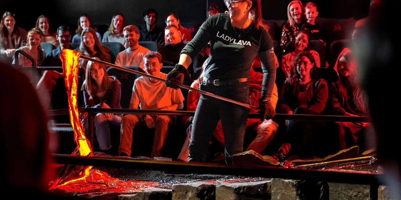 A host with a metal rod beside glowing molten lava in front of a seated audience