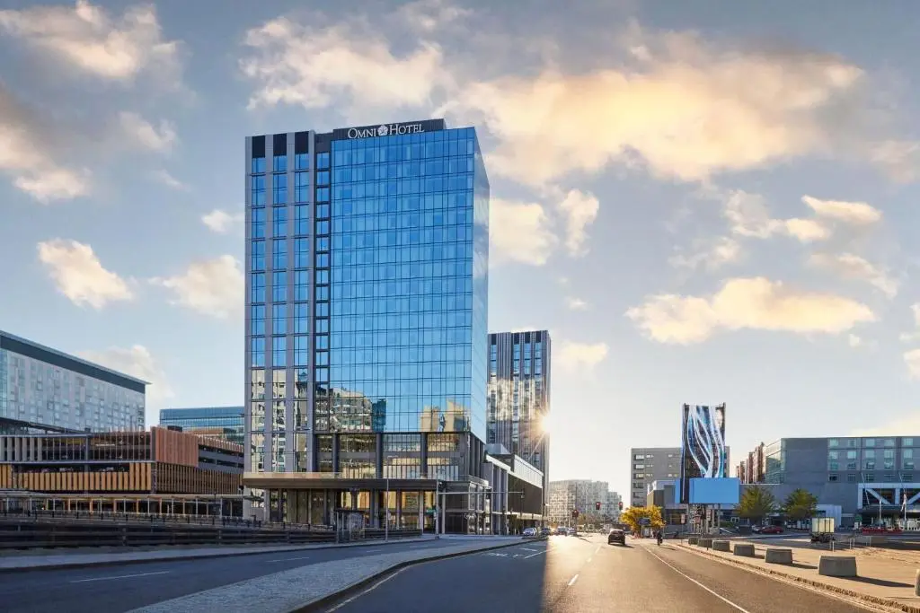 The glass tower of the Omni Boston Hotel at the Seaport on Summer Street, catching late sun under a blue sky with clouds
