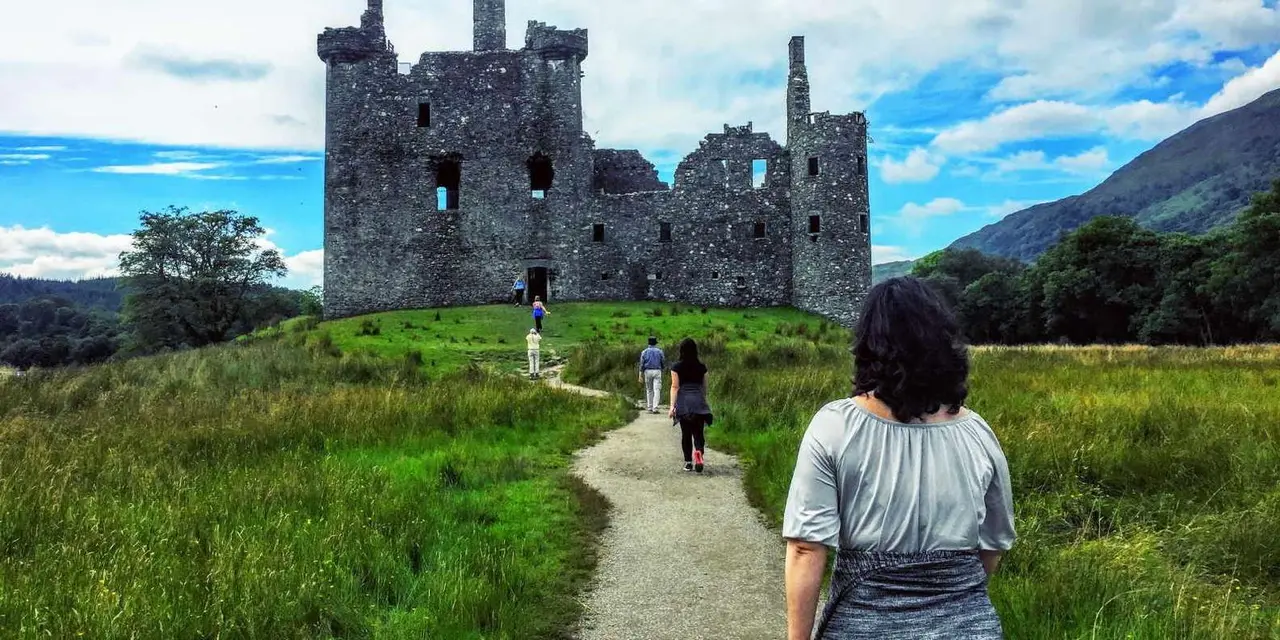 Kilchurn Castle's roofless stone ruin on a grassy rise, with visitors walking up the gravel path toward it