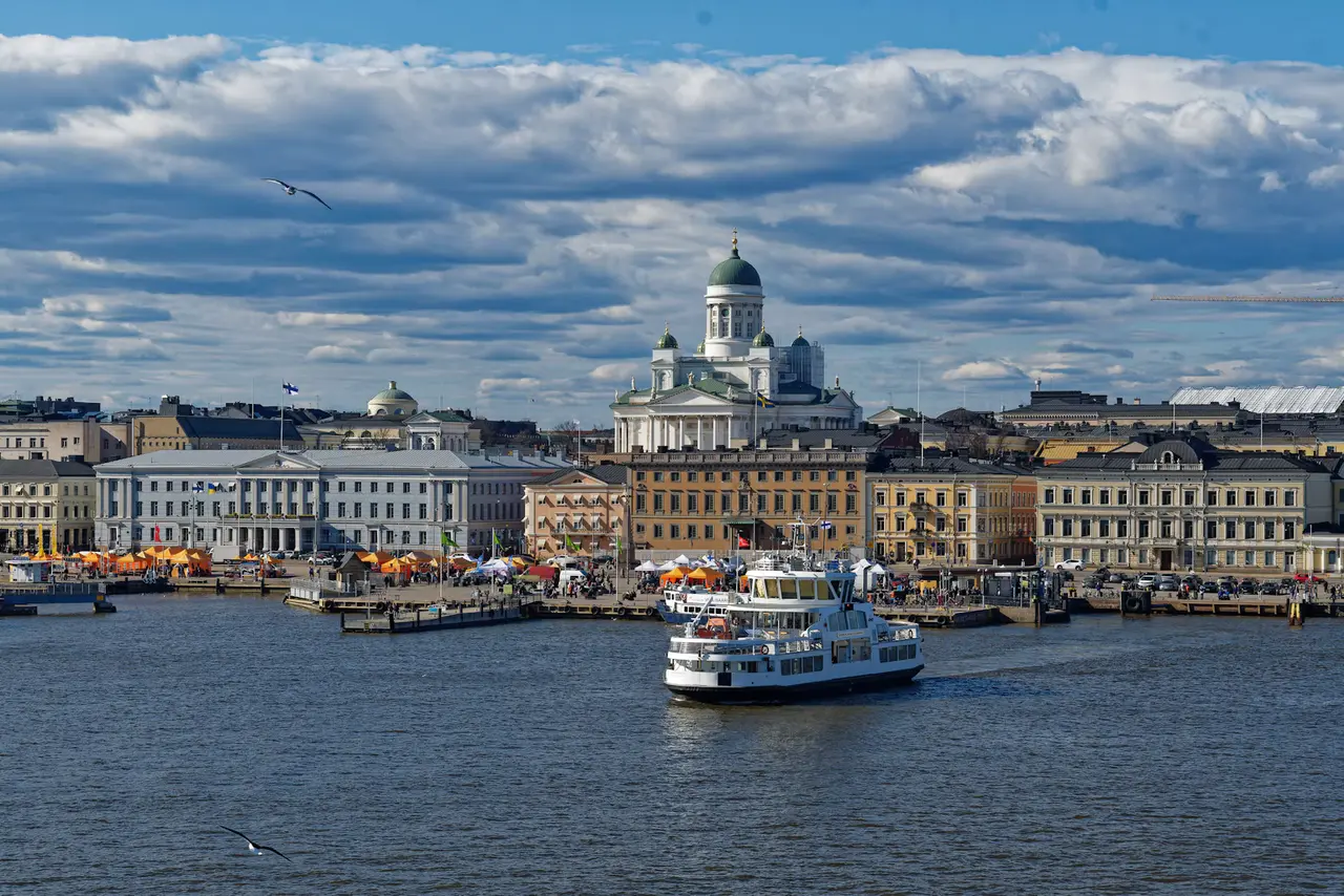 A white harbour ferry crosses the water in front of Helsinki Cathedral, the Market Square tents and the pastel waterfront buildings under a blue sky with big clouds
