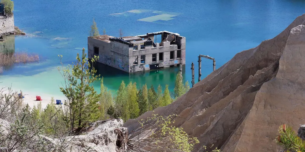 A ruined concrete building rising from bright blue-green quarry water, with a pale sandy cliff in the foreground