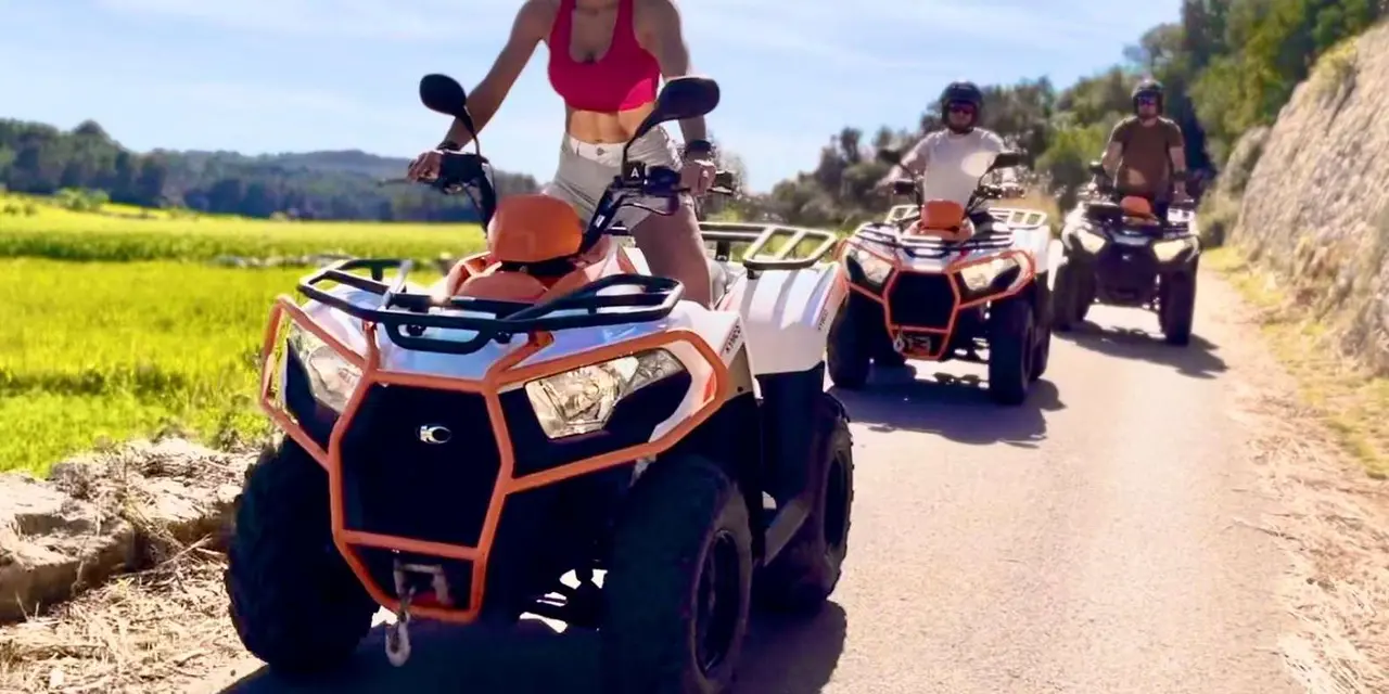 Riders on orange and white quad bikes on a country road with green fields behind