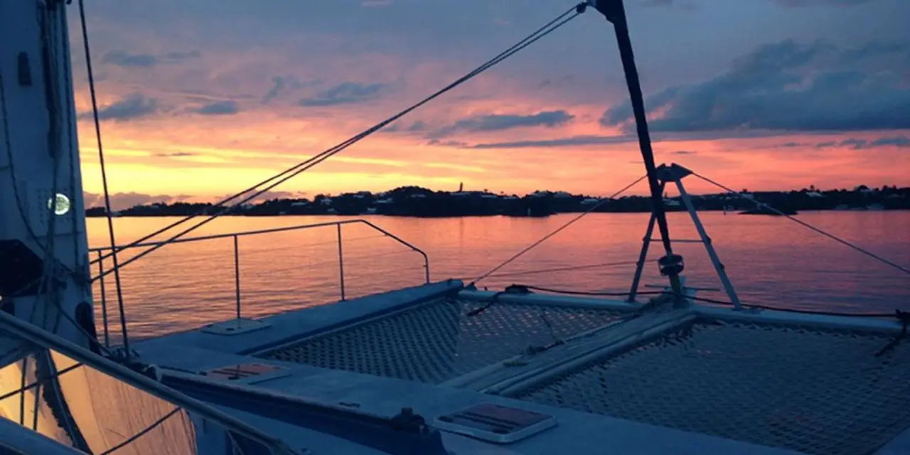 A catamaran's deck and rigging silhouetted against an orange and pink sunset over the water