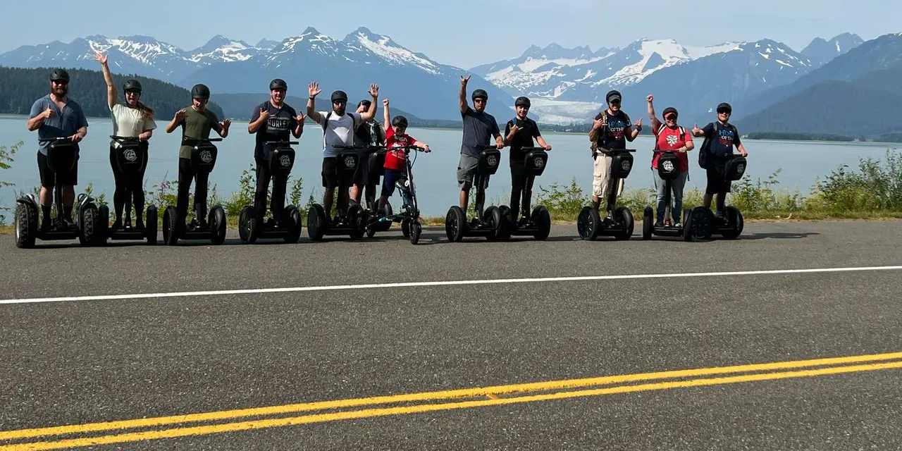 A line of helmeted riders on Segways with arms raised on a road, with calm water and snowy peaks with a glacier behind them