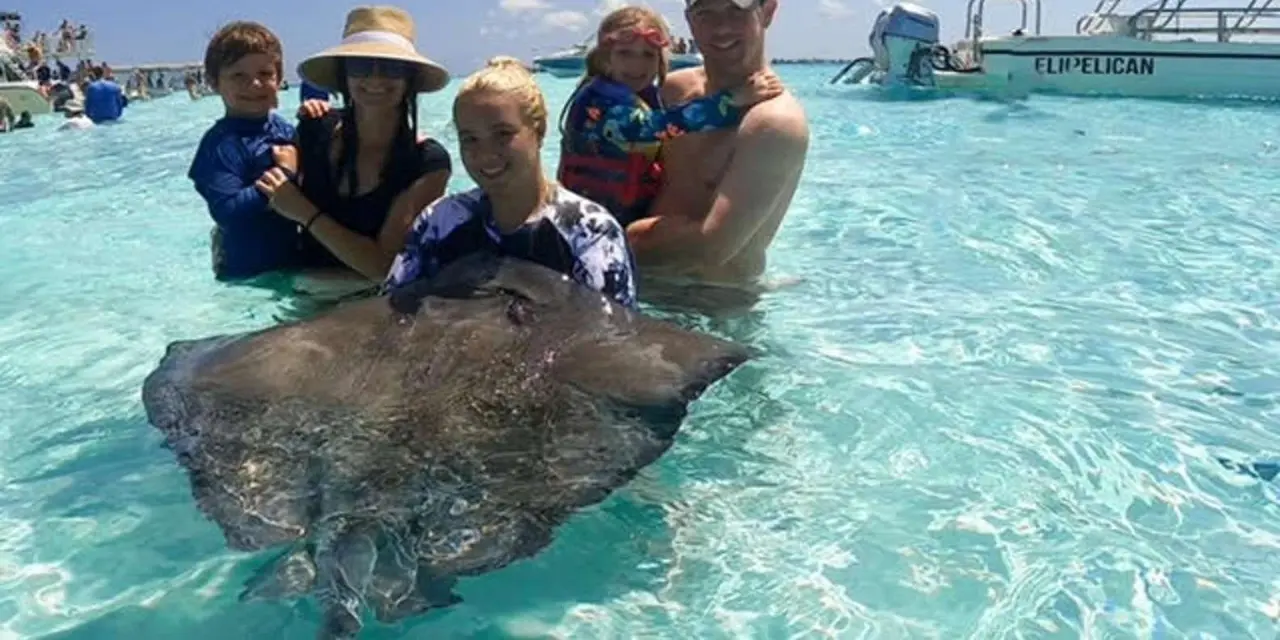 A family standing chest-deep in clear turquoise water at the Stingray City sandbar with a large stingray in front of them and a tour boat behind