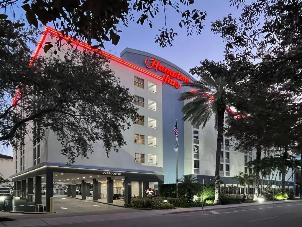 The Hampton Inn Coconut Grove's white tower with its red neon sign at dusk, framed by oak branches and palms
