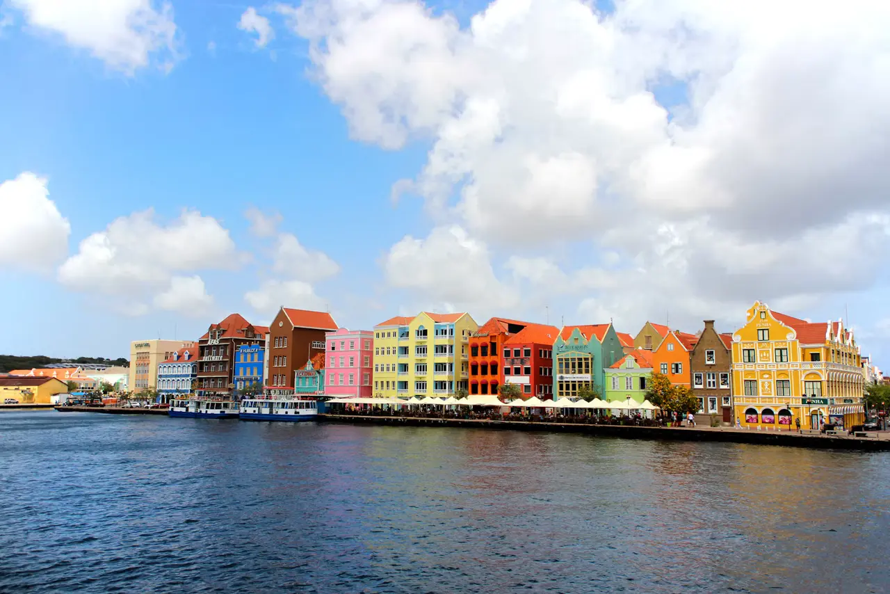 The pastel Dutch-colonial houses of Willemstad's Handelskade waterfront in Curaçao, seen across St. Anna Bay from the Queen Emma Bridge under a big blue sky with white clouds
