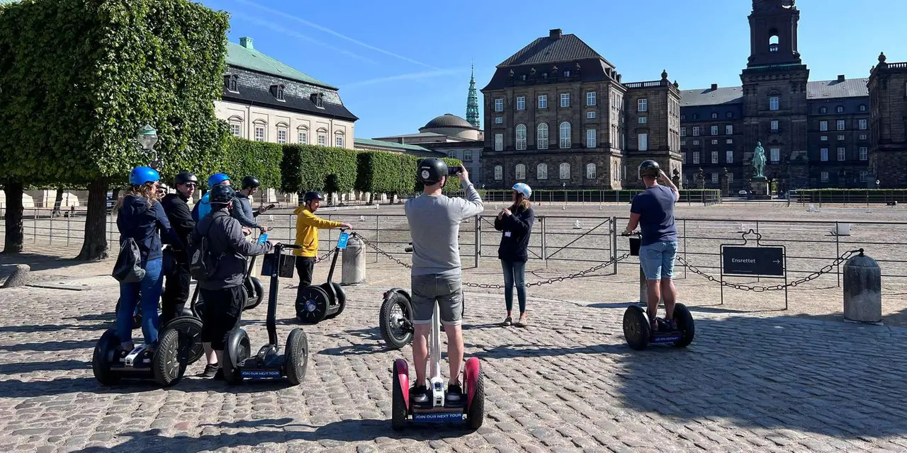 A group in helmets on Segways on cobbles, with a guide talking in front of the riding ground and the dark stone facade of Christiansborg Palace