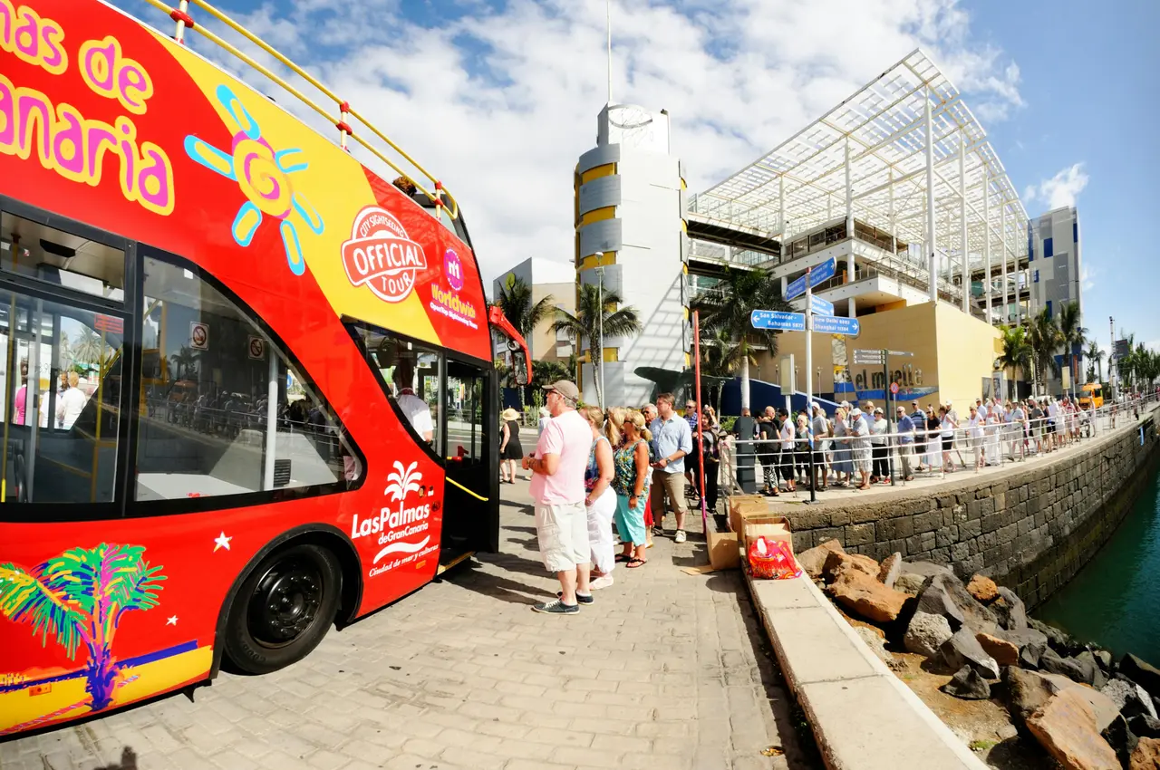 A red open-top double-decker bus with passengers queuing on the seafront beside a modern glass building