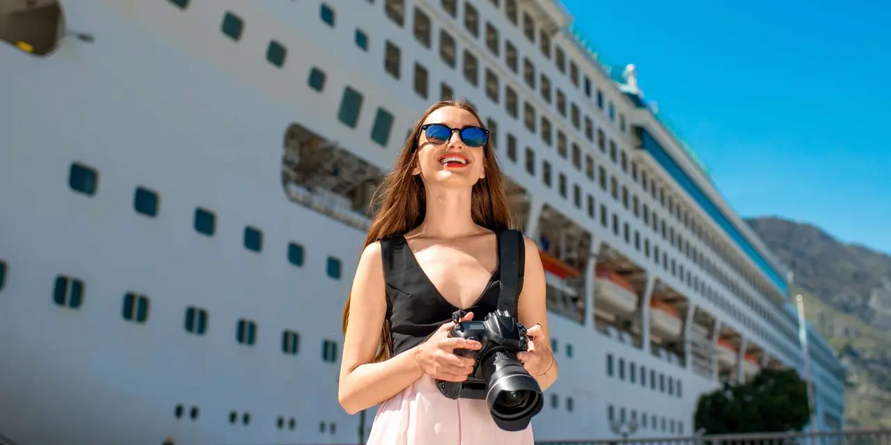 A smiling woman holding a camera on the quay beside a large white cruise ship under a blue sky