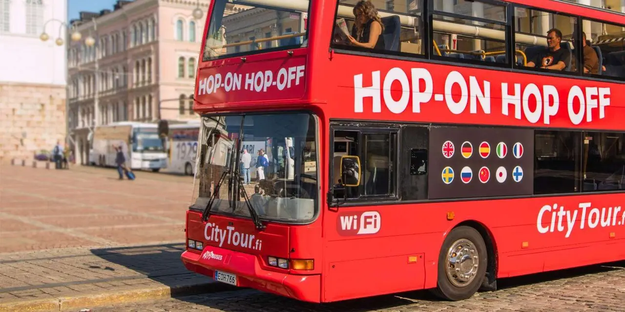 A red open-top double-decker bus marked Hop-On Hop-Off beside an old stone building, with other buses and pedestrians in the background