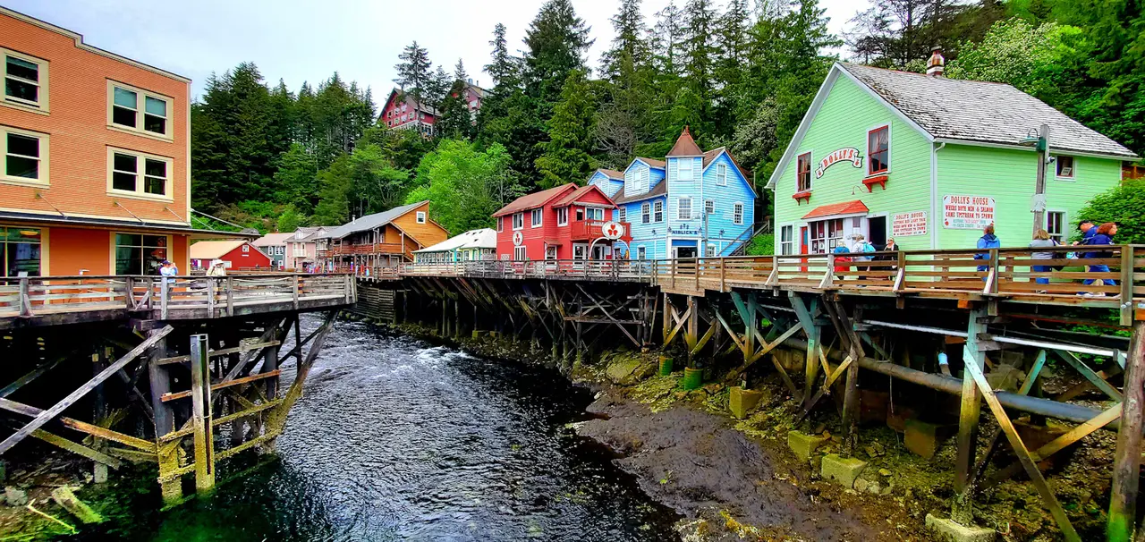 Creek Street in Ketchikan: brightly painted wooden houses, mint green, red, blue and orange, standing on pilings above a rocky creek, with tall spruce behind