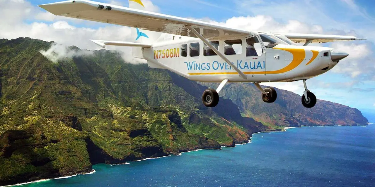 A white Wings Over Kauai plane flying above steep green ridges and the blue Na Pali coastline