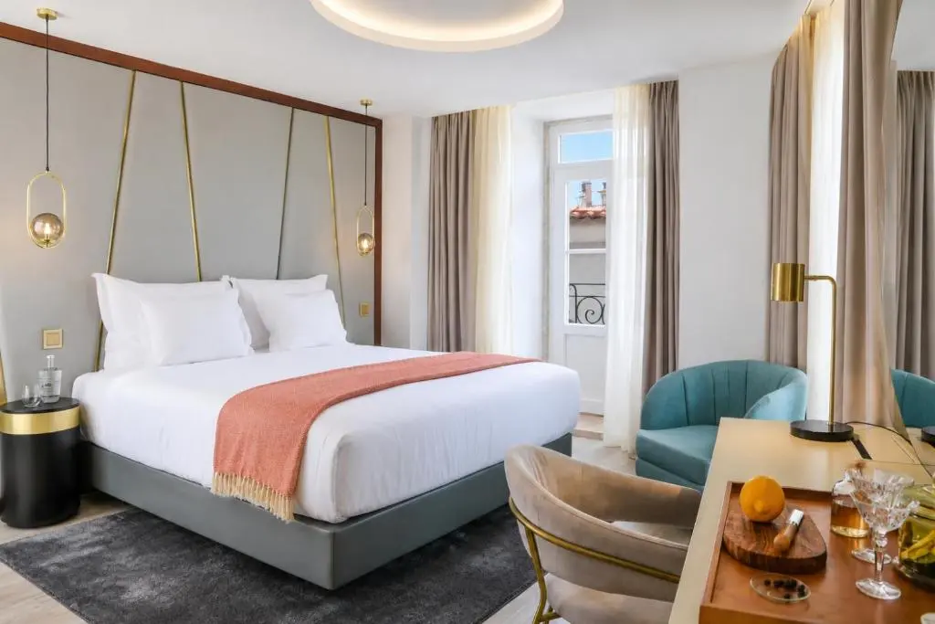 Guest room at Vincci Alfama with a double bed under a coral throw, brass wall lamps, a teal armchair and a tall window to a small balcony