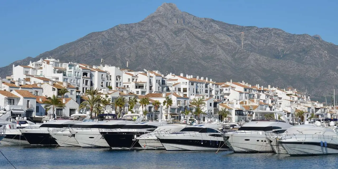 A white hillside town at the foot of a rocky mountain, with a marina full of boats in front