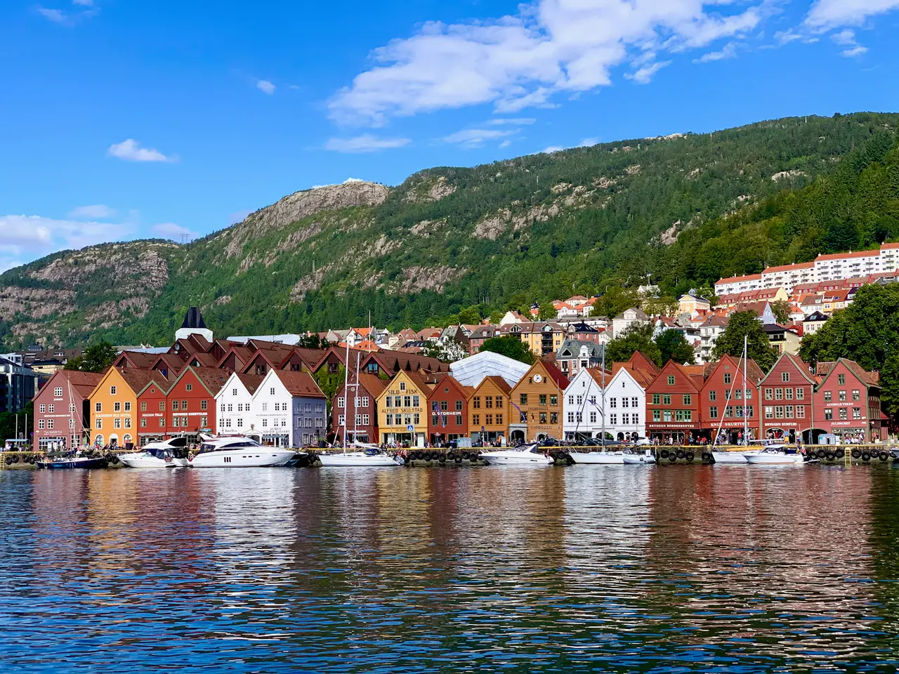 Bryggen's row of red, orange and white wooden wharf houses across Bergen's harbour on a sunny day, boats moored below and a green mountain behind under blue sky