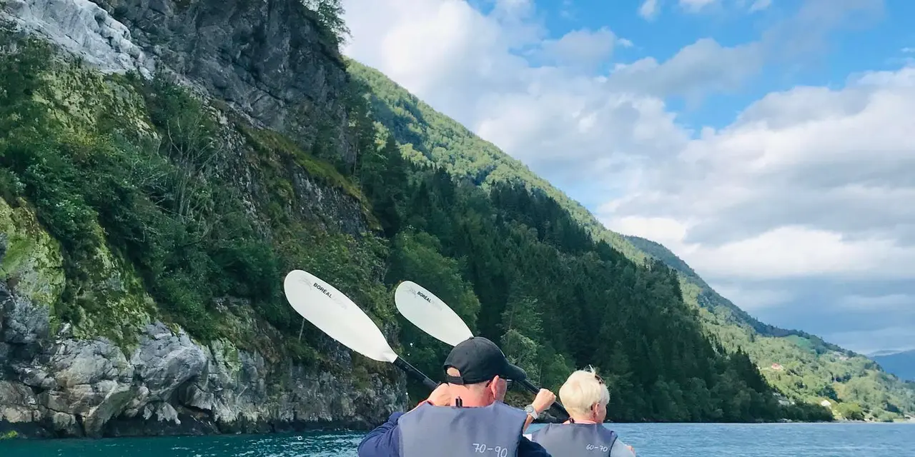 Two paddlers seen from behind with paddles raised, facing a wooded cliff and sky
