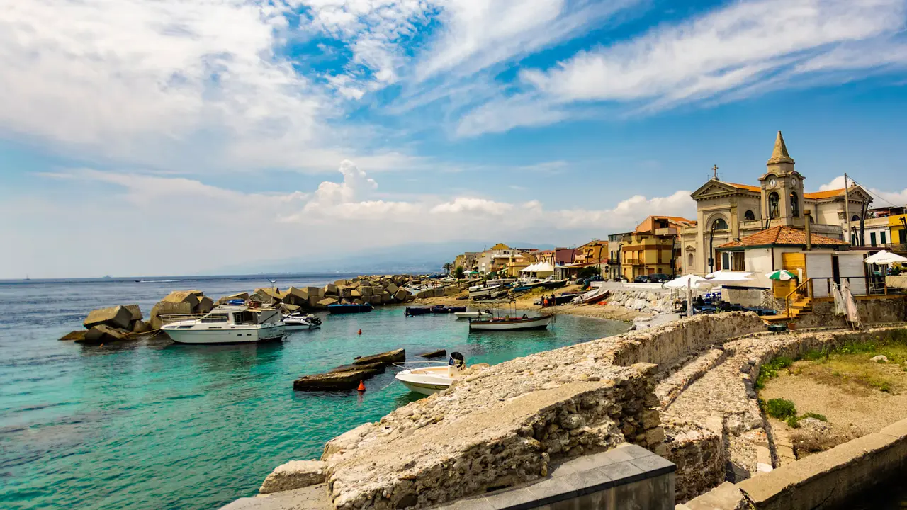 Small boats moored in clear turquoise water below a seafront church and pastel houses on Messina's coast, under a wide blue sky streaked with cloud