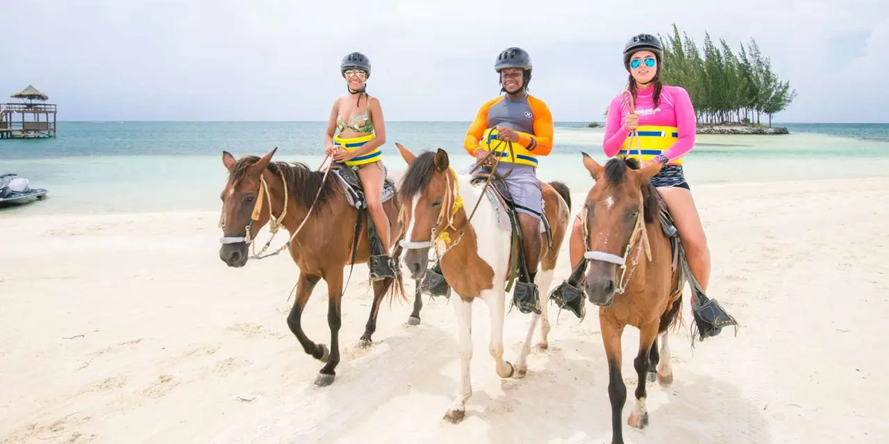 Three riders in helmets and life vests on horseback along a white-sand beach with turquoise water behind