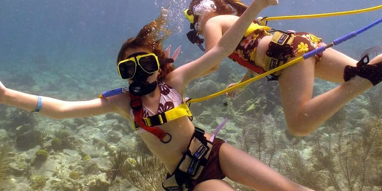 Two snorkel-masked swimmers underwater above a reef, breathing through yellow air hoses that run up to the surface