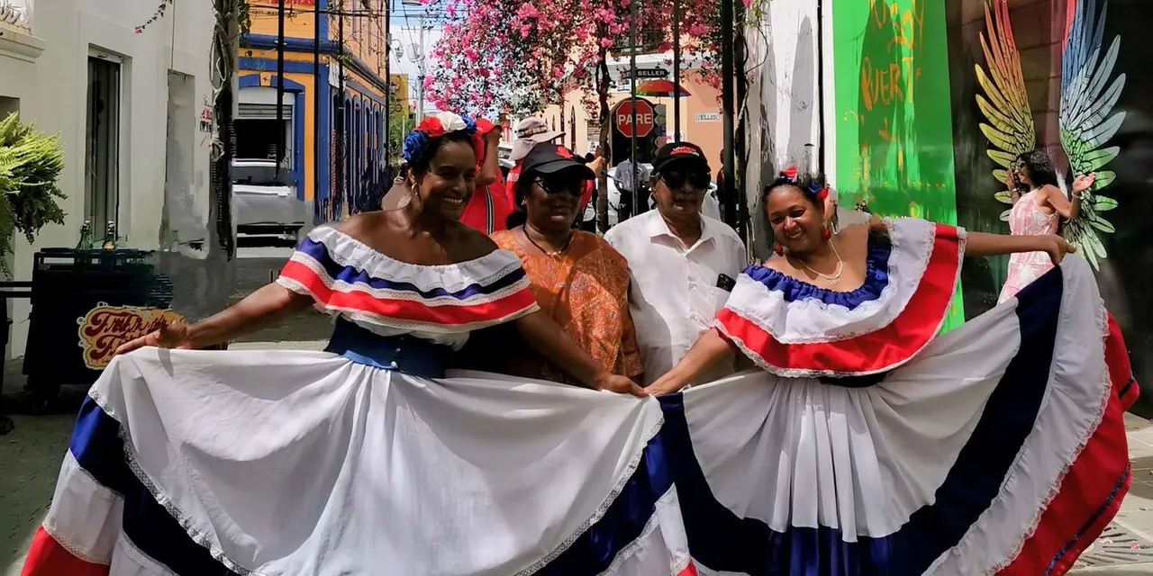 Dancers in white skirts with blue and red trim posing under colourful hanging umbrellas on a Puerto Plata street