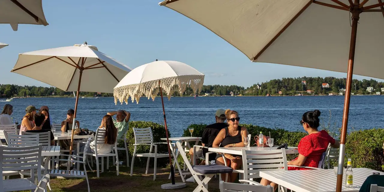 People at white café tables under parasols on a waterside lawn, with forested islands across the water