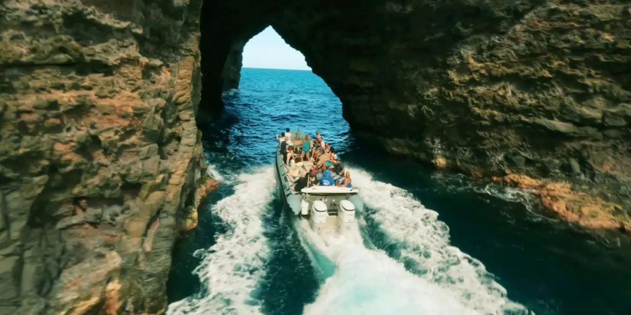 A Zodiac raft full of passengers speeding out through a rock arch of a sea cave into open blue water