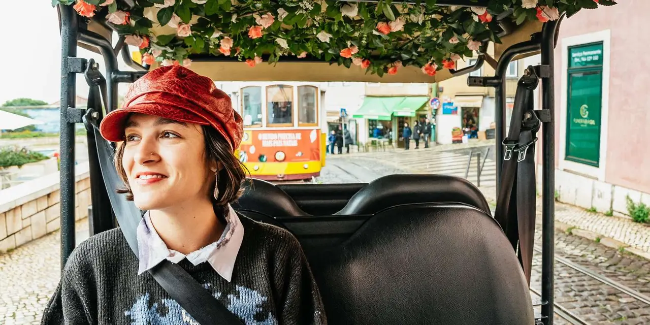 A smiling passenger in a red cap in the back of a tuk-tuk with a yellow tram passing behind