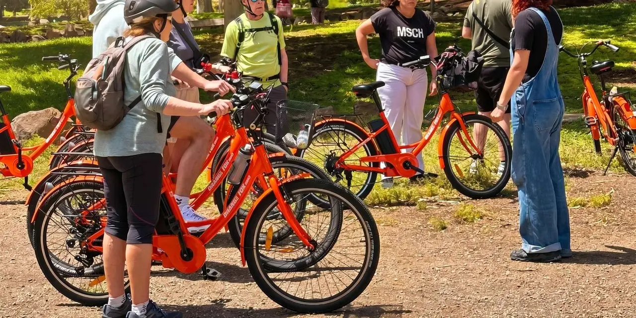 Visitors on orange rental bikes gathered on a gravel path beside grass, listening to a guide