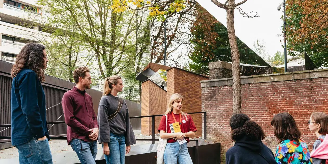 A guide in a red t-shirt holding a book about Anne Frank talks to a small group of visitors beside a brick memorial and autumn trees