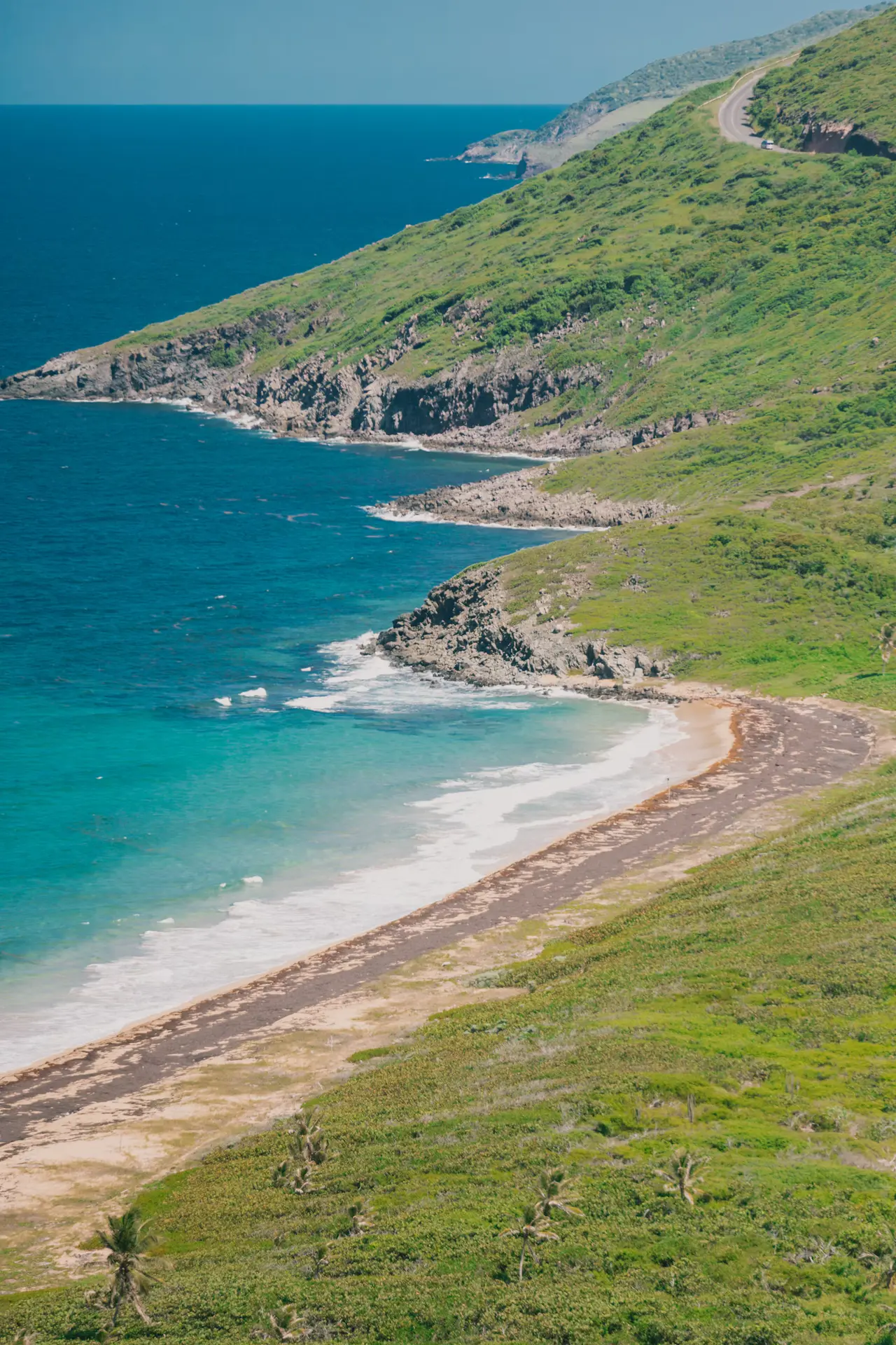 Turquoise surf breaking on a curved beach below steep green headlands on St. Kitts, a coast road winding along the ridge above