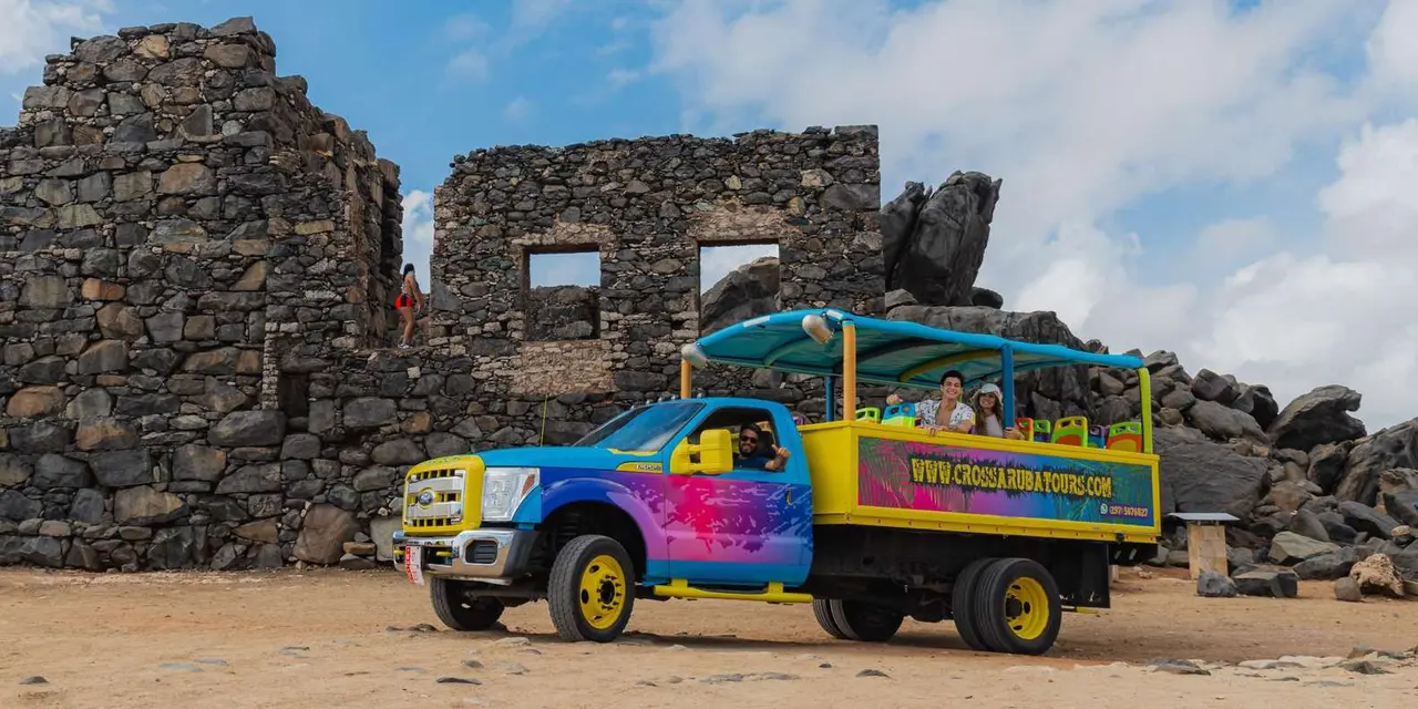 A colourful open-sided tour truck parked in front of the dark stone walls of the Bushiribana gold-mine ruins, with passengers aboard