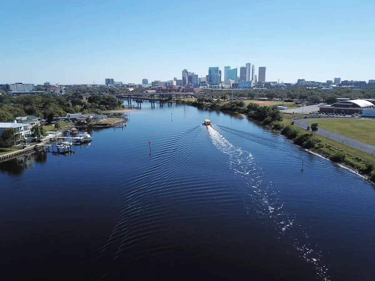 Aerial view down the Hillsborough River to the downtown Tampa skyline under a clear blue sky, a water taxi's wake trailing behind it