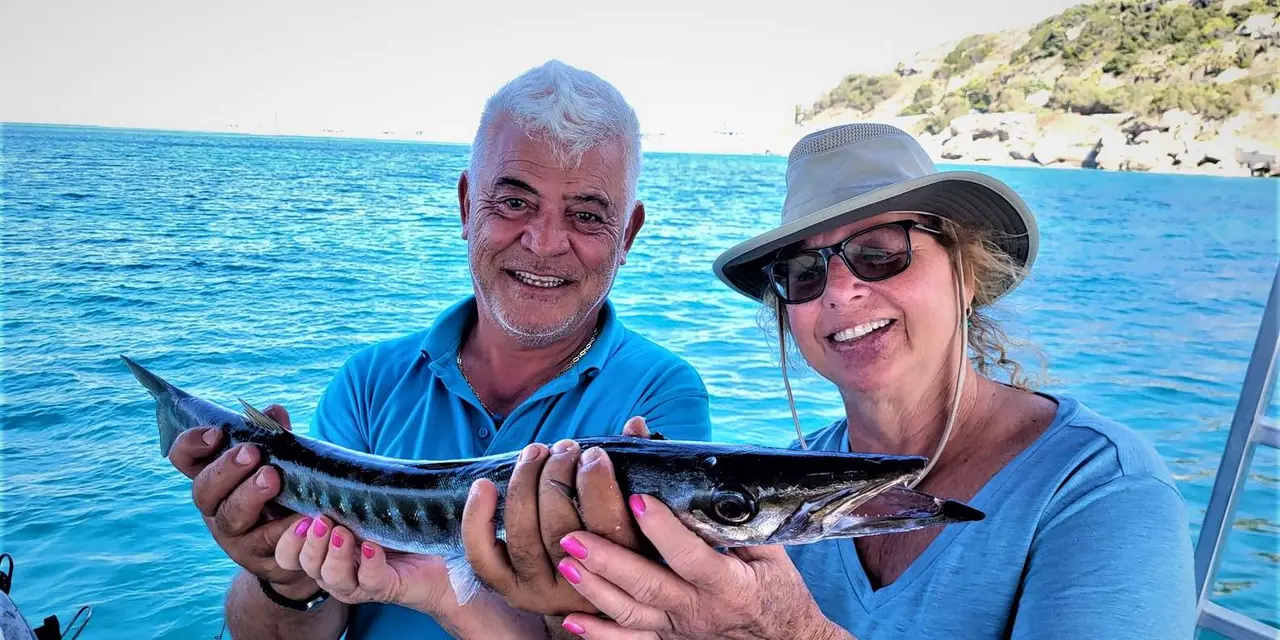 A man and a woman in sunglasses and a sun hat holding a long barracuda on a boat, blue sea and a rocky shore behind