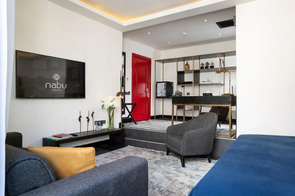 Suite at Nabu Hotel Karaköy with a grey sofa and armchair, a wall-mounted TV, a red door and a raised desk area