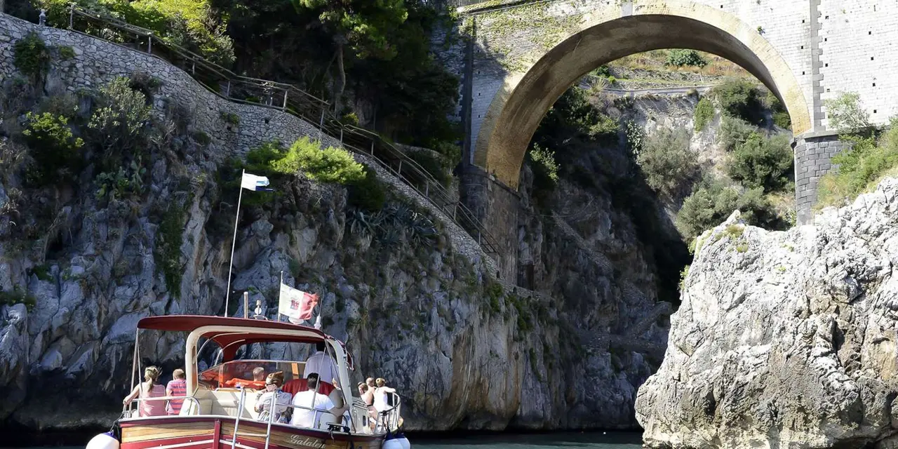 A stone arch bridge spanning a narrow rocky inlet on the Amalfi Coast, with a small boat in the water below