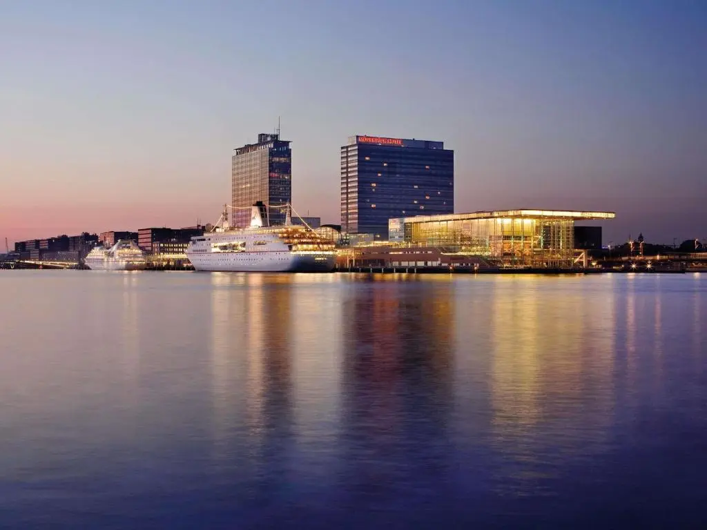 Mövenpick Hotel tower on the IJ at dusk beside the Muziekgebouw, with a white cruise ship moored at the quay in front