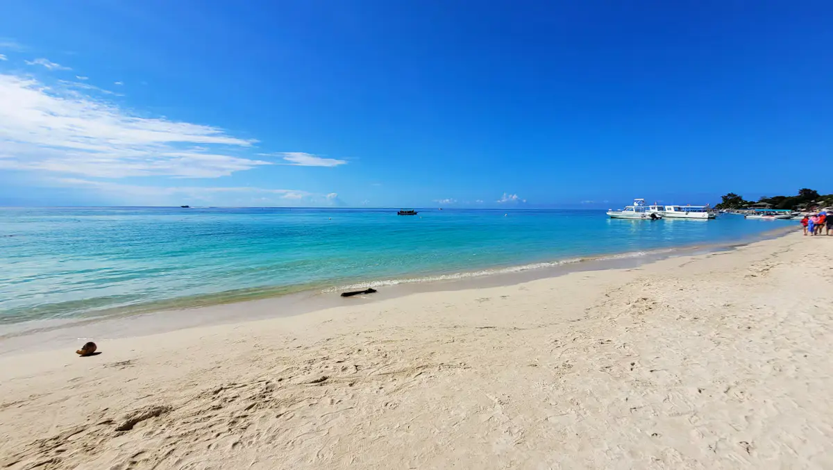 A long white-sand beach at West Bay, Roatán, with turquoise shallows, a few tour boats anchored at the far end and a deep blue sky with thin cloud
