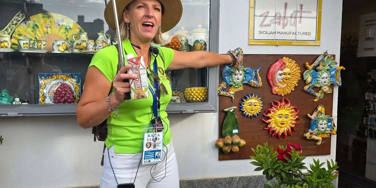 A smiling guide in a sun hat and lanyard pointing to a wall of painted ceramic suns in a Sicilian shop