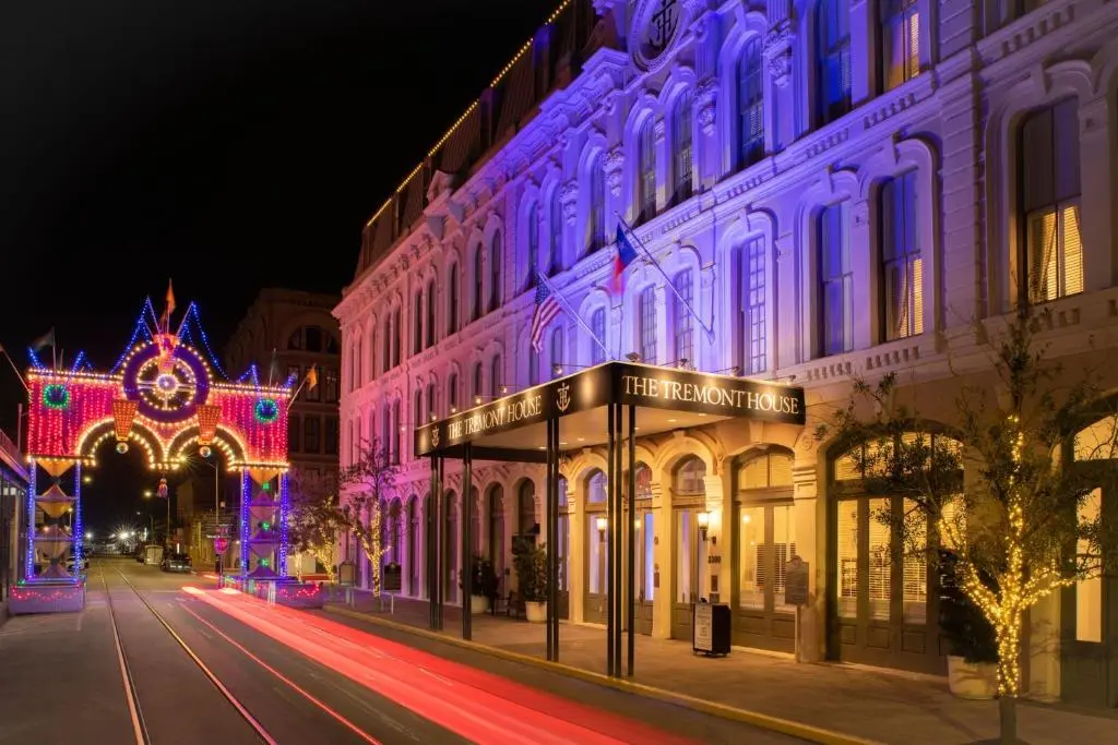 The Tremont House's lit-up Victorian facade and entrance canopy on a Galveston street at night, with a string-light archway beside it
