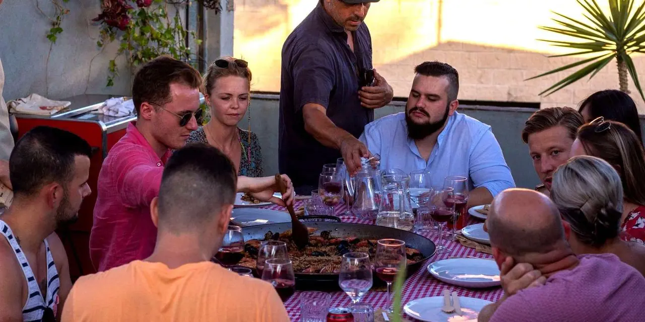 Guests around a long checked-cloth table on a sunlit rooftop, with a wide paella pan of seafood rice being served and glasses of red wine