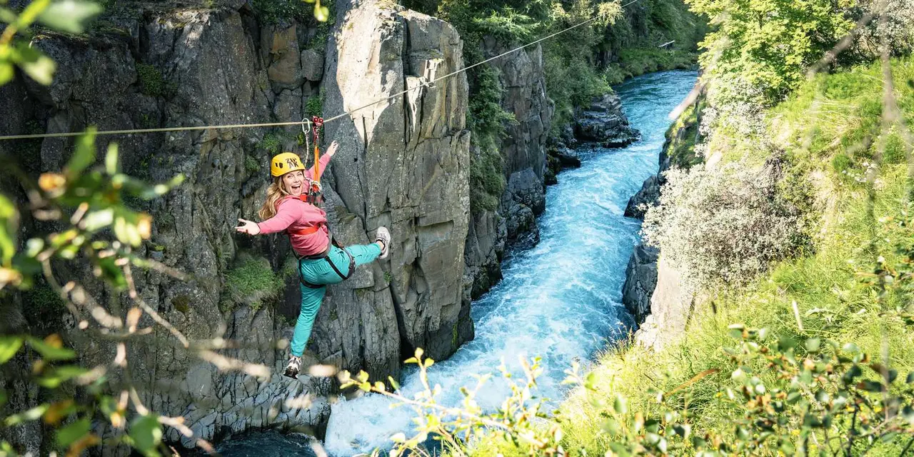 A smiling zipliner in a yellow helmet and pink top hanging mid-line above a rushing blue river in a rocky canyon